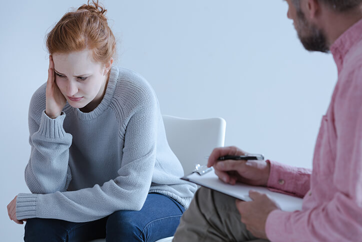 Young red headed woman with depression wearing a sweater is talking with a male therapist wearing a pink shirt.